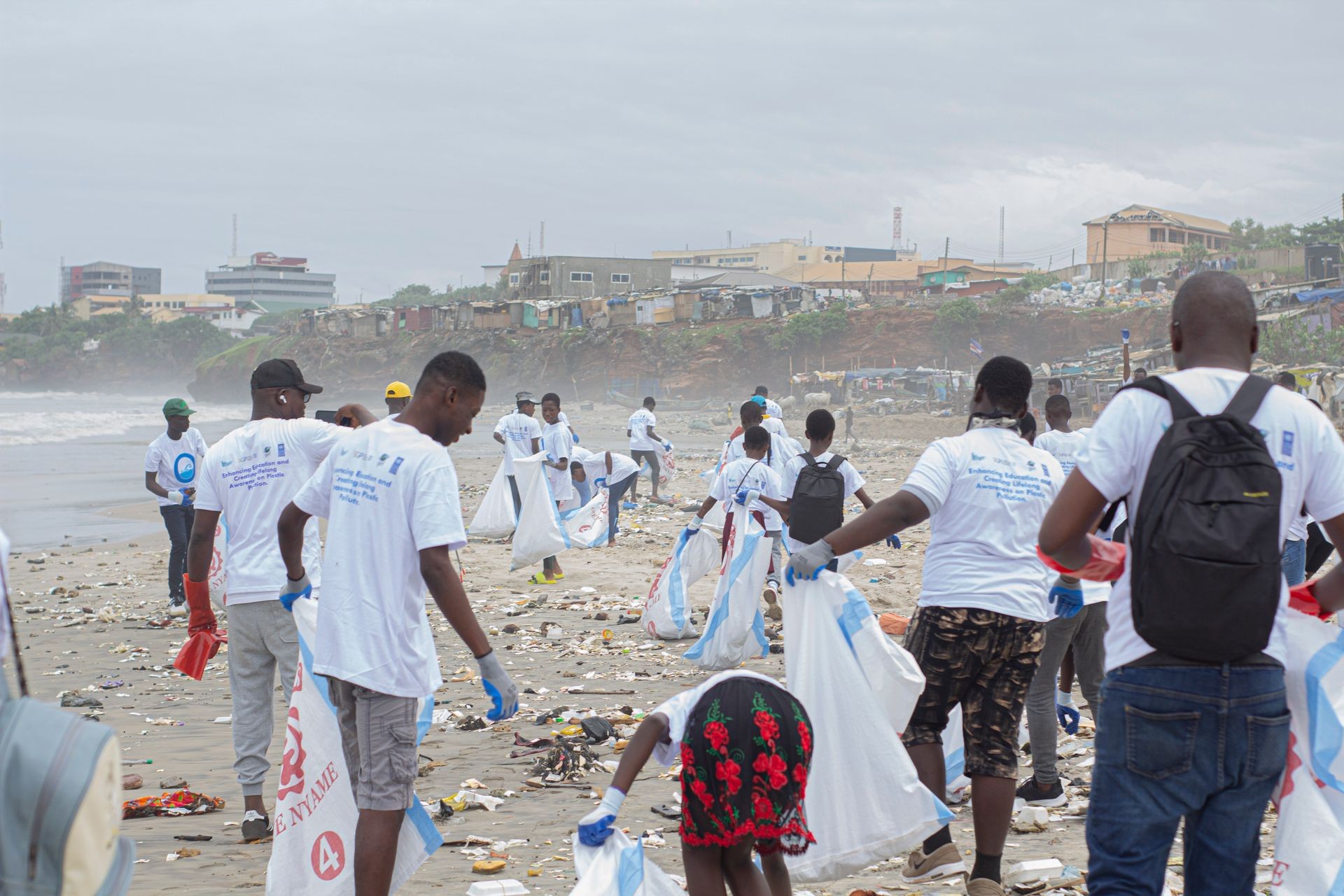 Group of volunteers cleaning a beach littered with trash, holding large bags and wearing white shirts.