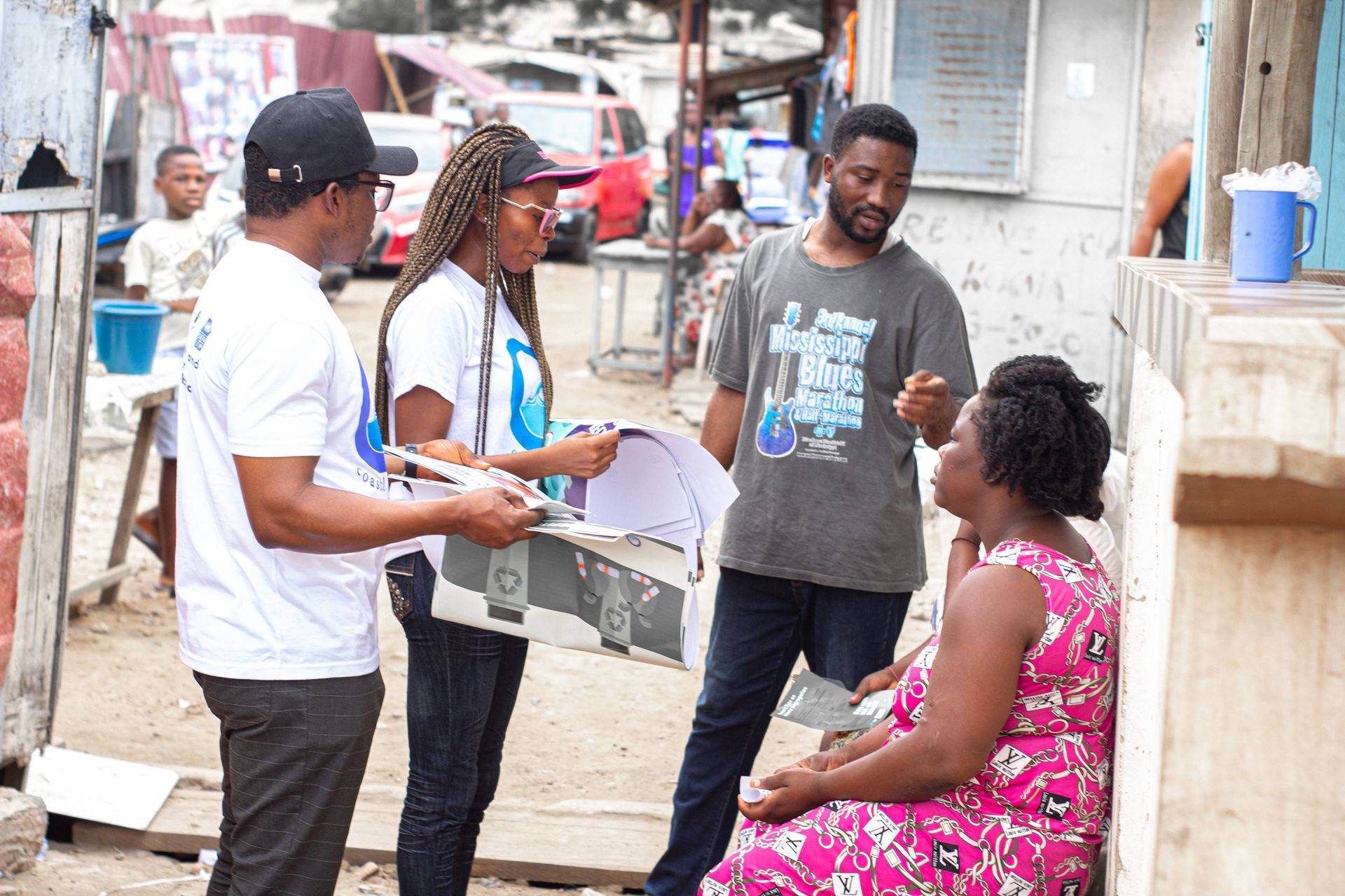 Group of people engaging in conversation outdoors, with one person seated and others standing holding papers.